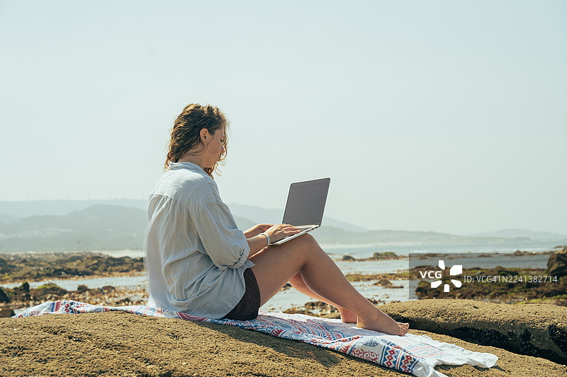 年轻女子在海边岩石上远程使用笔记本电脑工作——海滩上的数字游民生活方式图片素材