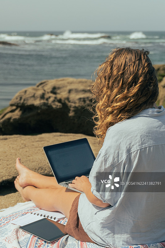年轻女子在海边岩石上用笔记本电脑远程工作——海滩上的数字游民生活方式图片素材