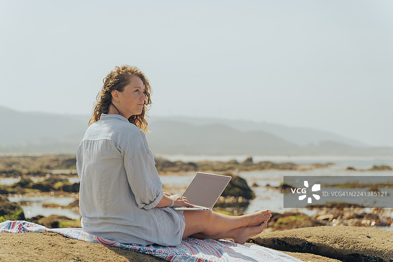 年轻女子在海边岩石上远程使用笔记本电脑工作——海滩上的数字游民生活方式图片素材