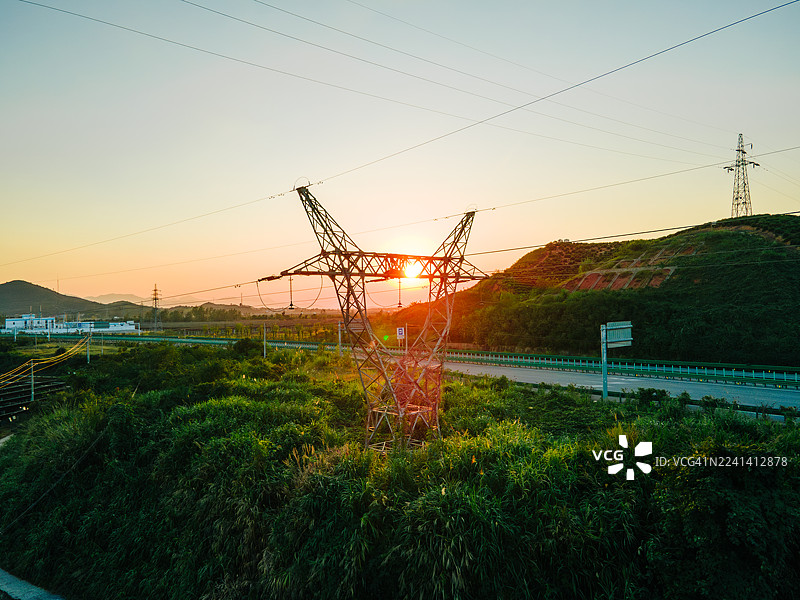 夕阳下，高压输电线路和铁塔横跨大地，在温暖的橙色天空下形成剪影，突显了电力基础设施和能源输送。图片素材