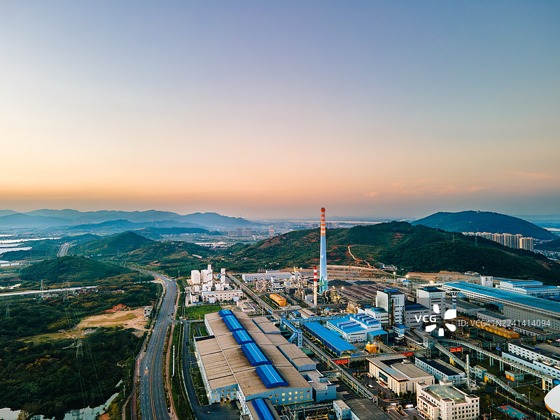 航拍工业工厂，有烟囱和石材加工厂，靠近高速公路，远处环绕着群山，在自然日光下，突显了工业运营、交通和景观的融合。图片素材