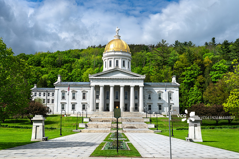佛蒙特州议会大厦在蒙彼利埃，标志性的金色圆顶在戏剧性的多云天空中显得格外醒目，周围环绕着郁郁葱葱的绿树和维护良好的草坪。图片素材