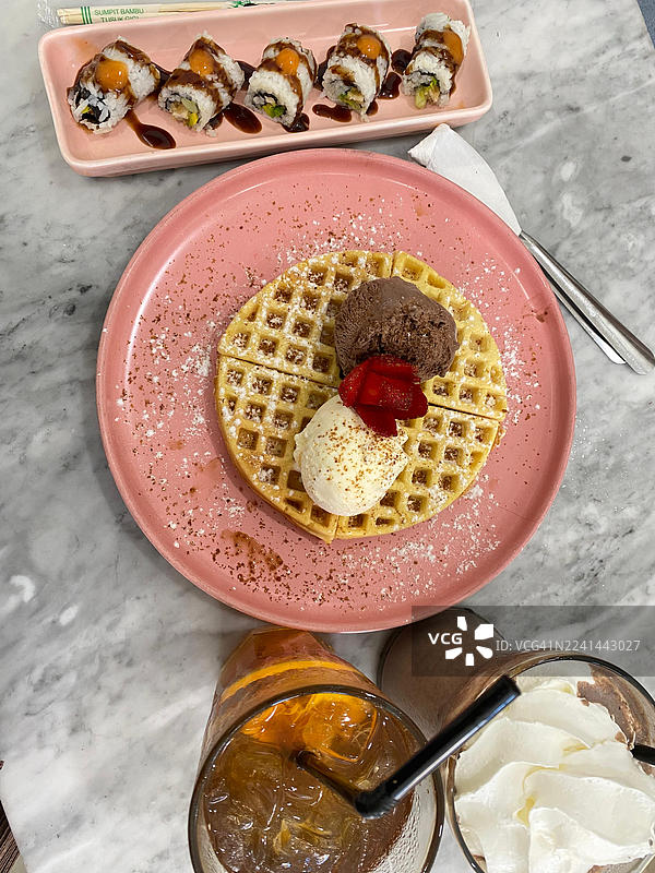 大理石桌上摆放着美味的华夫饼、冰淇淋和寿司，旁边还有饮品，俯视角度拍摄图片素材