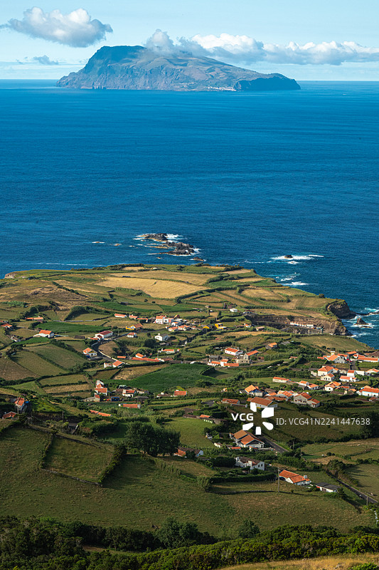 从高处俯瞰葡萄牙科尔武岛的海滨村庄，那里有梯田、广阔的蓝色大海和远处的岛屿，天空晴朗。图片素材