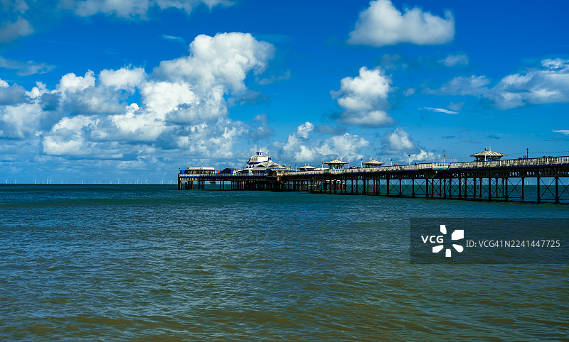 英国兰迪德诺，晴朗的蓝天和蓬松的白云下，长长的码头一直延伸到海里，远方还能看到海上风力涡轮机，风景优美。图片素材