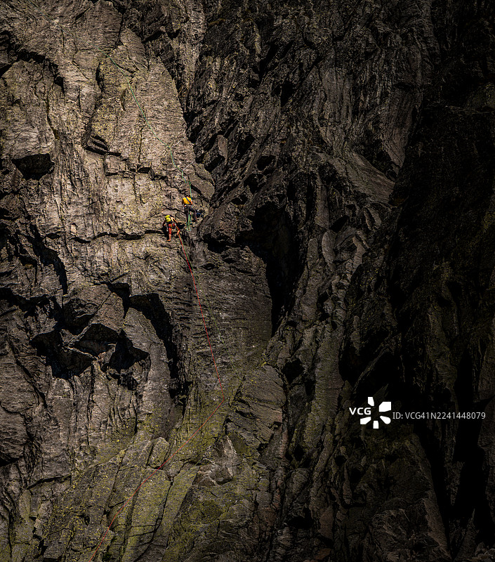 两名攀岩者一同攀登崎岖、阴影笼罩的山壁，他们系着绳索，共同迎接挑战，确保安全。图片素材