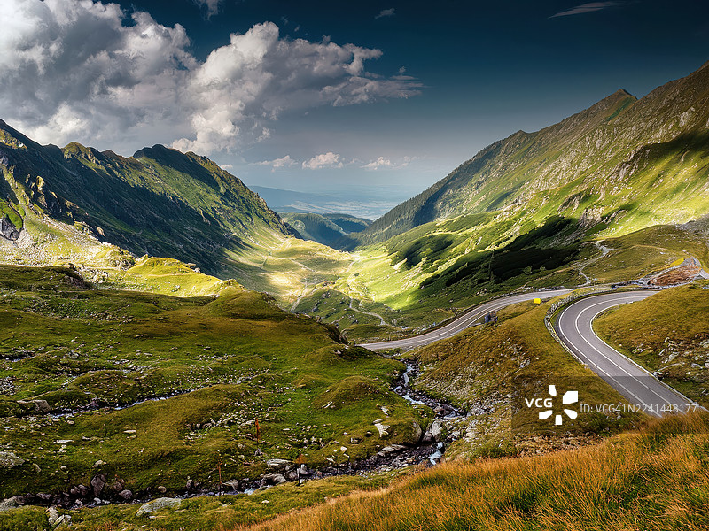 罗马尼亚，多云的天空下，蜿蜒的山路穿过郁郁葱葱的绿色山谷图片素材