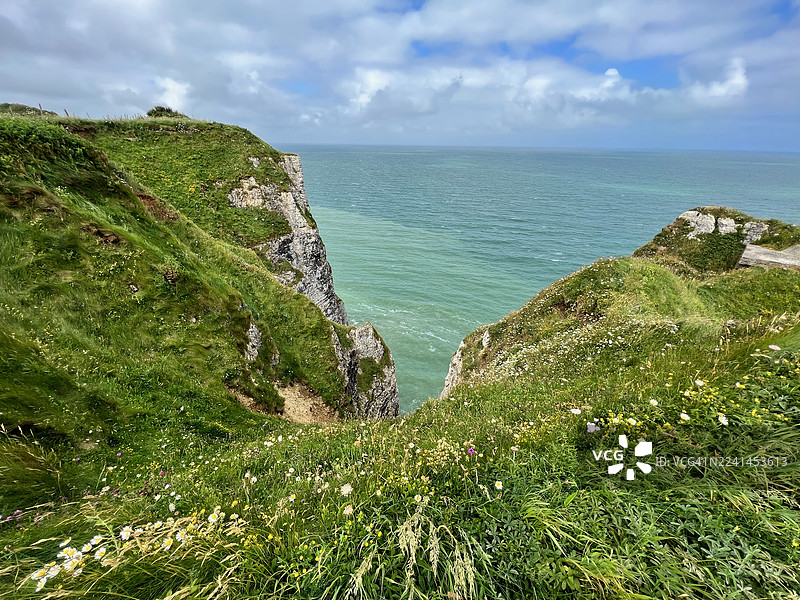 多云天空下，戏剧性的海岸悬崖俯瞰着绿松石般的大海，前景是郁郁葱葱的绿草和野花，法国图片素材