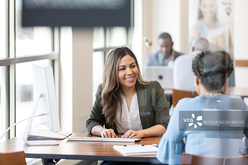一位职业女性在办公桌前与同事交谈时面带微笑。图片素材