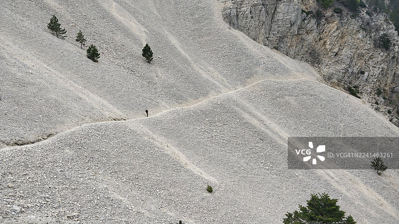 从高处俯瞰，一人正行走在蜿蜒的山路上，穿过一片广阔的、有稀疏树木的岩石山地景观，地点为法国的旺图山。图片素材