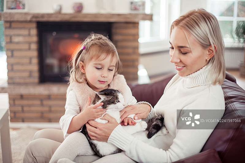 一位美丽的母亲、她的小女儿和一只猫正坐在壁炉旁的沙发上。家庭居家室内场景，温馨舒适。图片素材