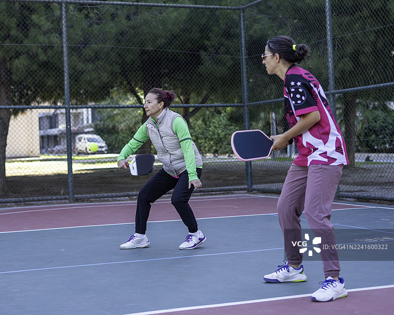 两位女性匹克球运动员正在硬地比赛，其中一位球员正在打截击球，球悬在空中，匹克球静止。图片素材