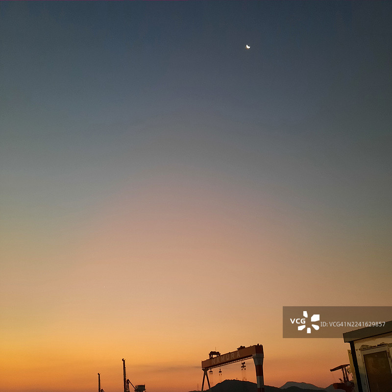 日本伊万里，日落时分，晴朗的天空中挂着一弯新月，远景是工业起重机和山脉的风景图片素材