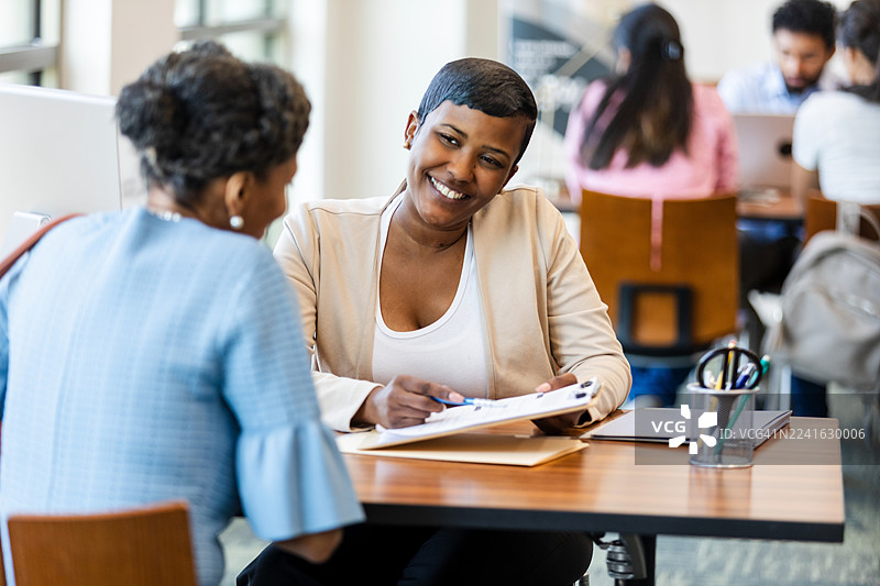 两位女性同事在办公室的办公桌旁，微笑着讨论文件。图片素材