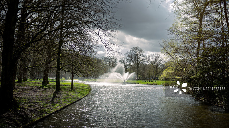 荷兰阿珀尔多伦公园的风景，喷泉位于蜿蜒的河流中，背景是戏剧性的多云天空，河岸边绿树成荫。图片素材