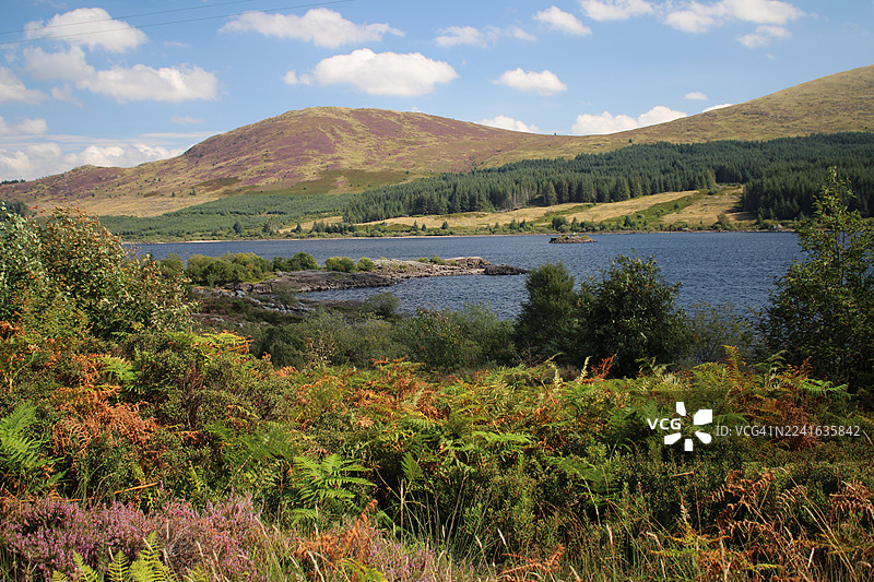 苏格兰达尔梅灵顿洛赫多恩城堡附近宁静湖泊的风景，湖泊被连绵起伏的山丘和茂密的植被环绕，天空湛蓝，点缀着几朵白云图片素材