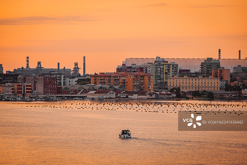 日落时分，一座拥有工业元素的海滨城市景观，水面上还有一艘船，意大利塔兰托图片素材
