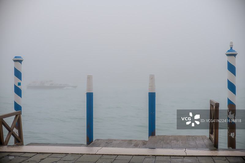 薄雾弥漫的早晨，一艘传统的威尼斯水上出租车正驶近一个带有蓝白条纹系船柱的码头，地点：意大利，威尼斯。图片素材