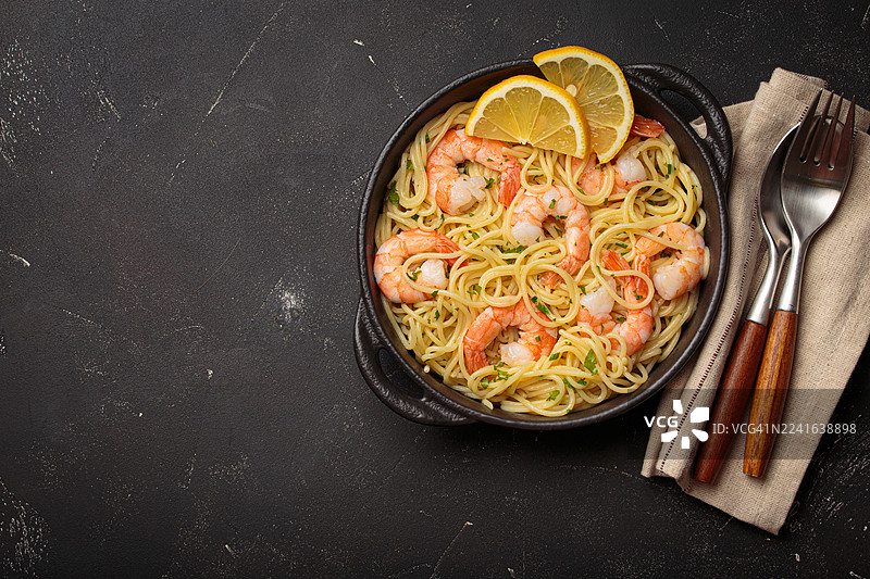 黑色碗中盛放的美味意大利虾面，配以柠檬片，置于深色质朴的桌面上，旁边摆放着餐具，俯视角度拍摄。图片素材