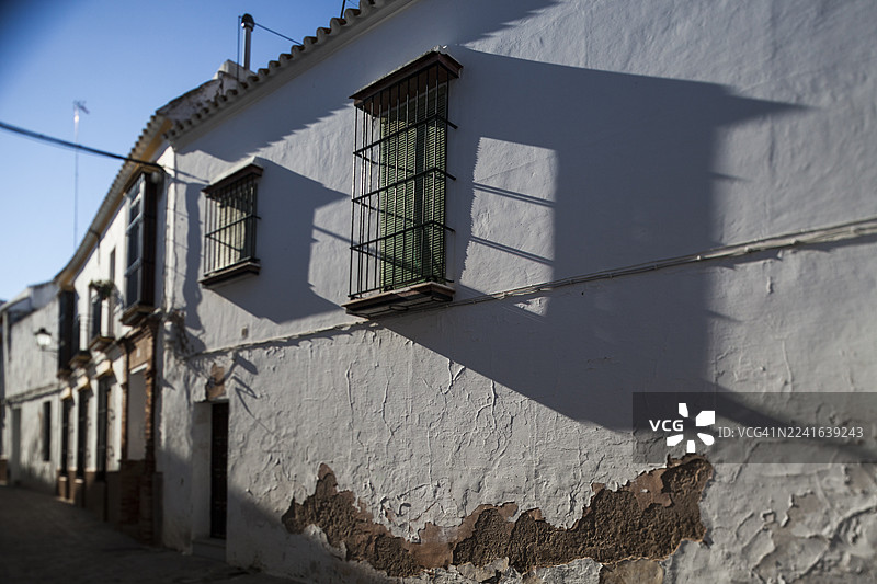 低角度拍摄的住宅楼，外墙白色油漆剥落，窗户装有铁栅栏，背景是晴朗的蓝天，投下长长的影子，地点：西班牙卡尔莫纳图片素材