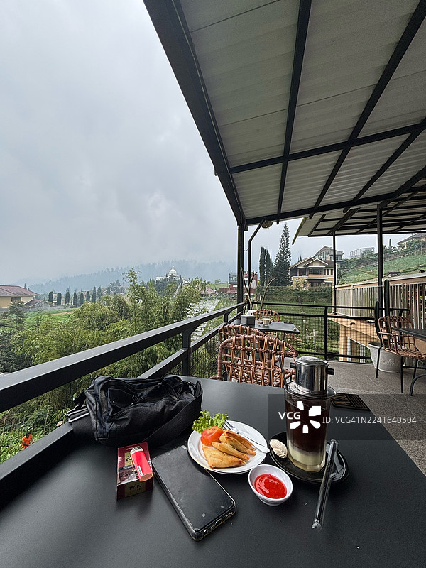 从阳台咖啡馆的高角度视角看餐桌上的食物和饮料，背景是风景如画的山脉和天空。图片素材