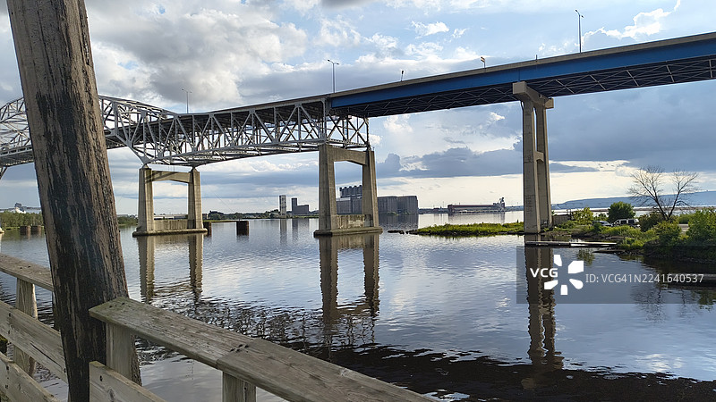 一座桥横跨宁静的河流，在戏剧性的多云天空下，远处是工业建筑。图片素材