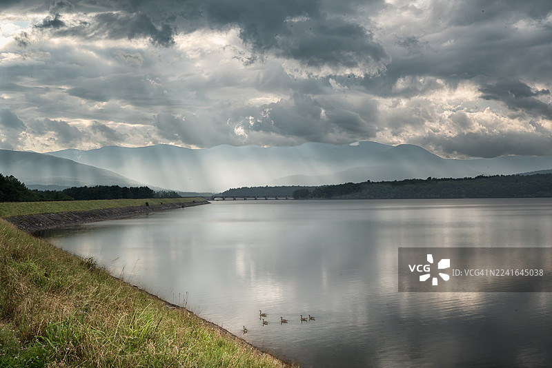 暴风雨云层中阳光穿透，远山、桥梁和宁静湖泊的戏剧性风景，橄榄，美国，纽约，阿什坎水库图片素材