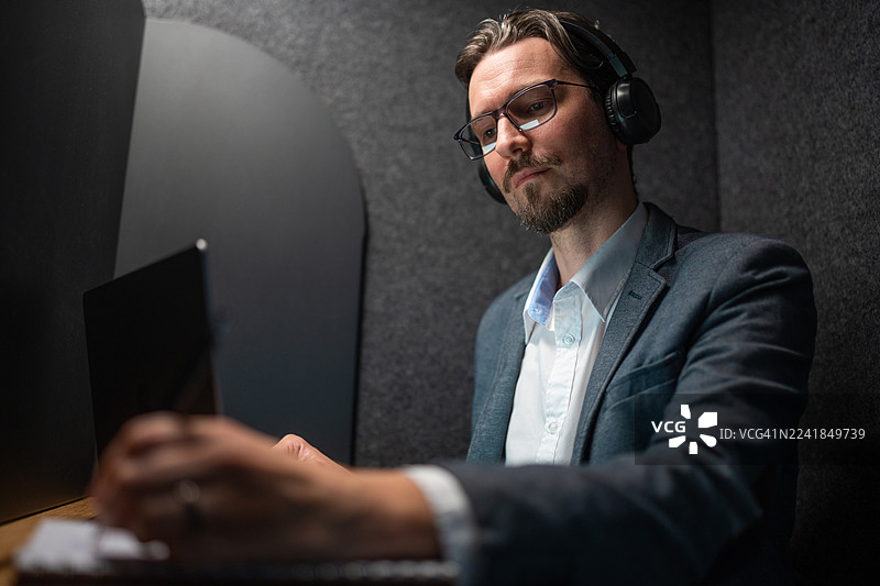 戴着耳机在独立隔间里使用平板电脑工作的沉思男人图片素材