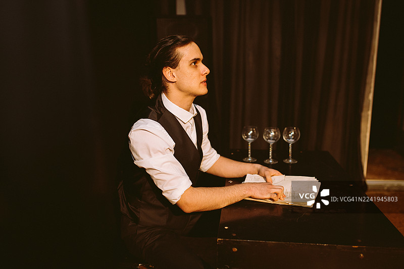 一名穿着背心的年轻男子坐在舞台的桌子旁，正在研读一出戏剧的剧本。图片素材