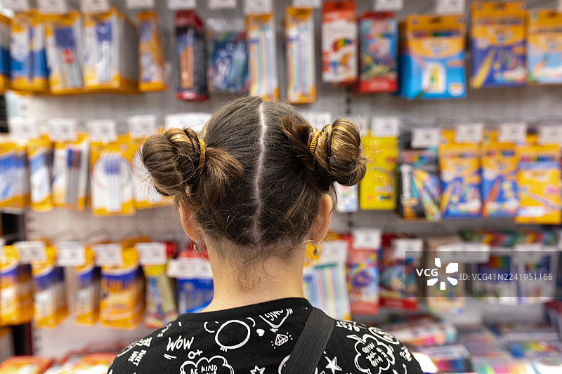 梳着双丸子头的女孩在色彩缤纷的文具货架旁购物，货架上摆满了各式文具用品。图片素材