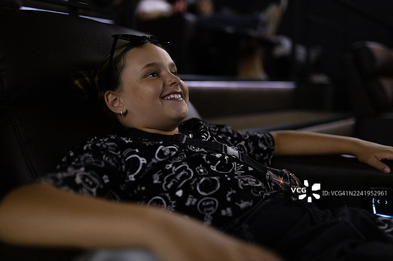 一个放松的女孩，微笑着斜倚在深色沙发上，头顶架着一副太阳镜，身处一个休闲舒适的环境中图片素材