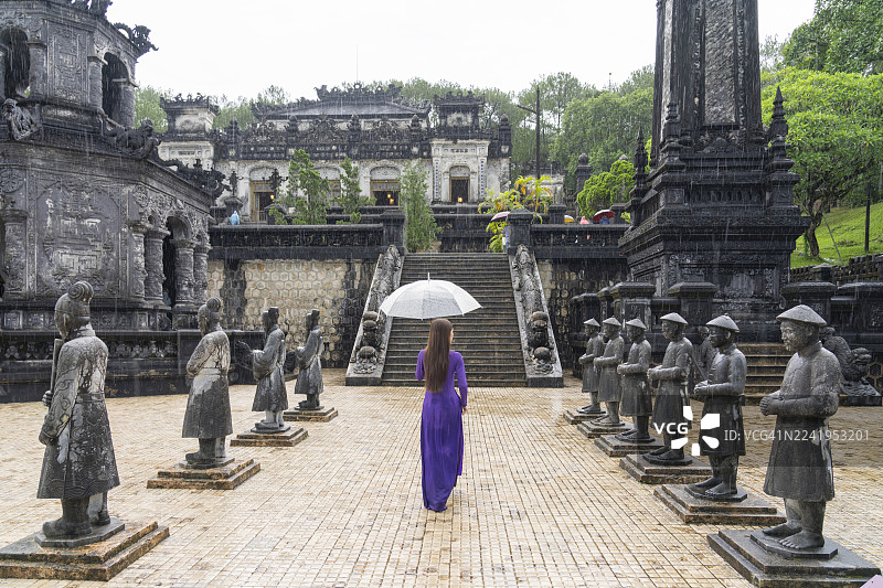 越南女子身穿紫色奥黛，撑伞走在顺化启定皇陵的雕像之间图片素材