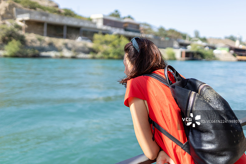 阳光明媚的一天，身穿红色衬衫的女子背着背包，倚靠在栏杆旁，旁边是蓝色河流。图片素材