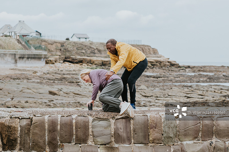 一对年长夫妇在码头上的滑稽而温馨的画面——男人试图扶起挣扎着站起来的女人，女人大笑着，男人也咯咯地笑了起来。图片素材