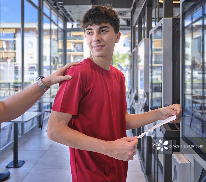 青少年在家长支持下使用自助服务终端点餐。他已经付款，手里拿着票。图片素材