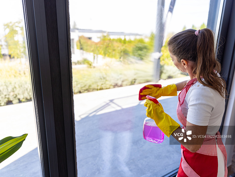 一位女士在家中明亮的场景下，用粉色喷壶和黄色手套清洁窗户图片素材