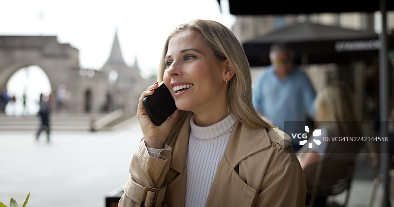 一位女子在咖啡馆放松地打电话并微笑，进行沟通、聊天或讨论。一位年轻女子在户外咖啡馆进行社交、联系和数字技术交流，进行对话、交谈和快乐的时光。图片素材