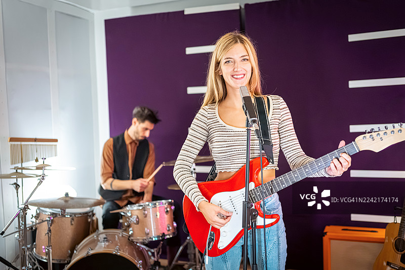 一位年轻女子在录音室里弹吉他，她身后的男性鼓手正在演奏，背景是紫色的墙壁图片素材