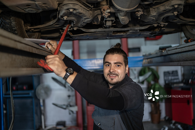 汽车修理店里，一位友好的机械师在举升机下用红色扳手修理汽车图片素材
