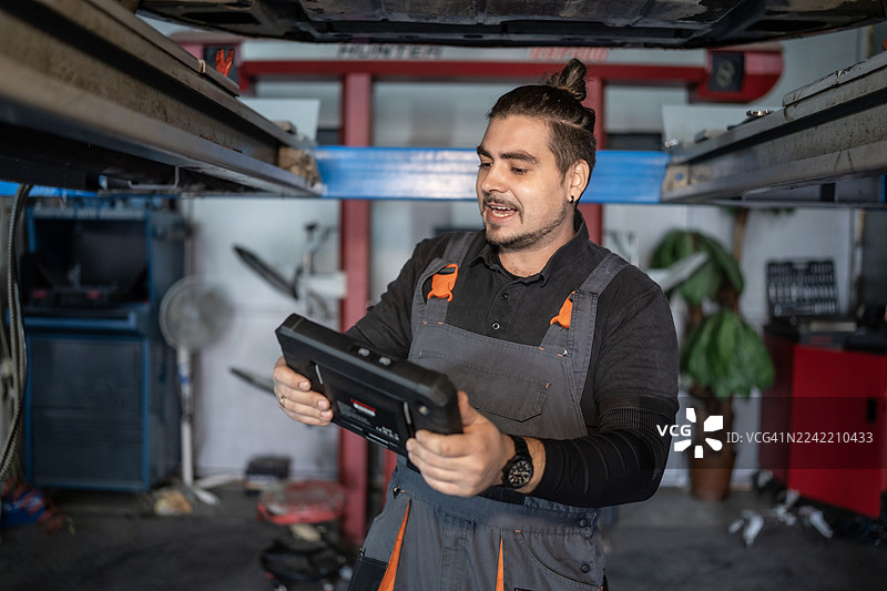 汽车修理技师穿着工装，在汽车维修店的车库升降机下使用诊断工具。图片素材