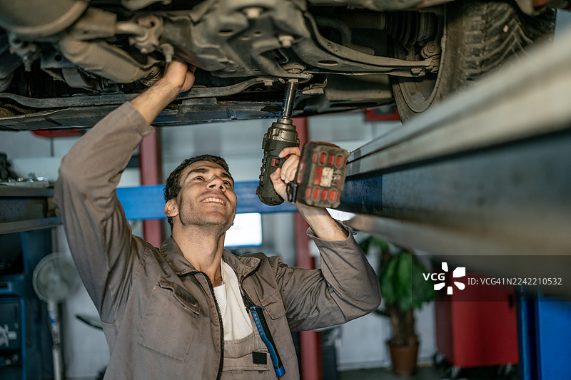 汽车修理工在汽车修理厂的升降车下使用钻头拧紧车身底部的部件图片素材