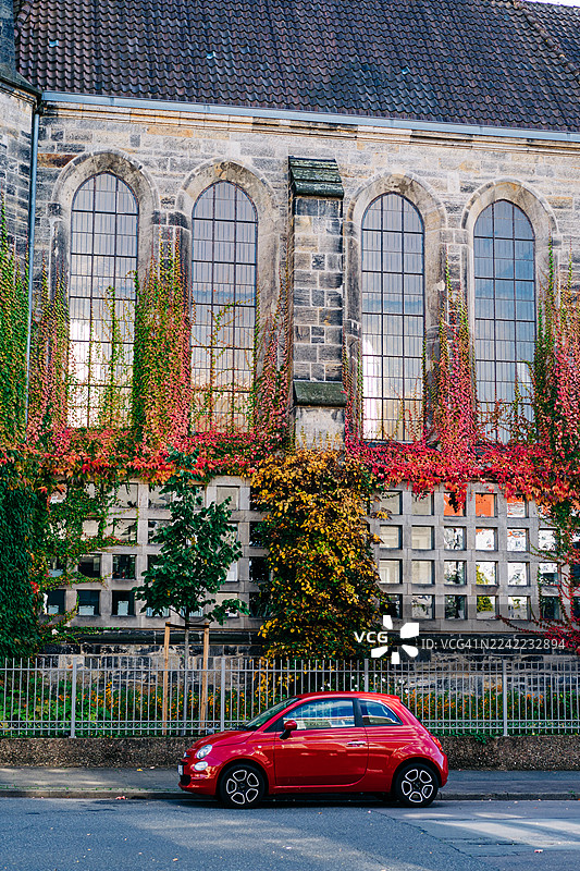 红色的紧凑型汽车停在爬满常春藤的石头建筑前，建筑有拱形窗户，位于城市街道上。图片素材