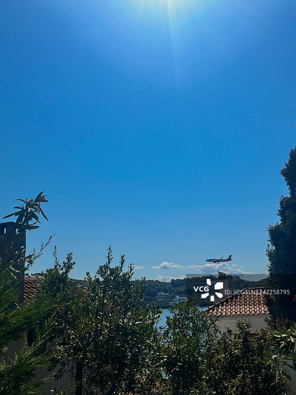 从低角度拍摄，一架飞机飞越海滨小镇上空，那里有郁郁葱葱的绿植和传统的瓦片屋顶，天空湛蓝明亮，地点是希腊的斯基亚索斯。图片素材