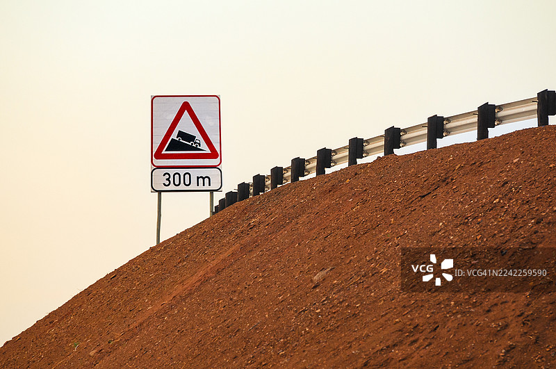 低角度拍摄的指示牌，标明卡车需要陡坡下降，背景是晴朗的天空，前景是护栏和泥土斜坡图片素材