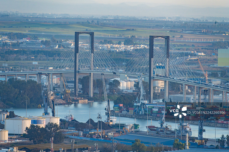 俯视一座现代斜拉桥跨越河流，前景是繁忙的港口和工业景观，背景是绿色的田野，地点：西班牙塞维利亚图片素材