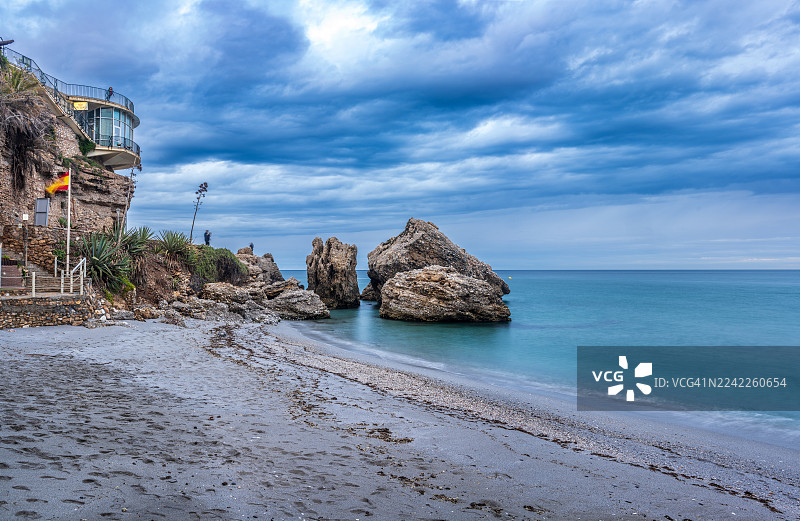 西班牙，悬崖上坐落着一座建筑，俯瞰着宁静的大海，海边是布满岩石的沙滩，天空阴沉多云，充满戏剧性。图片素材