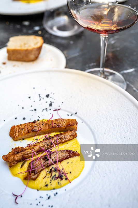白色盘子里盛放着美味的菜肴，有鲜嫩的肉或鱼、黄色酱汁和嫩苗，旁边配有面包和葡萄酒。图片素材