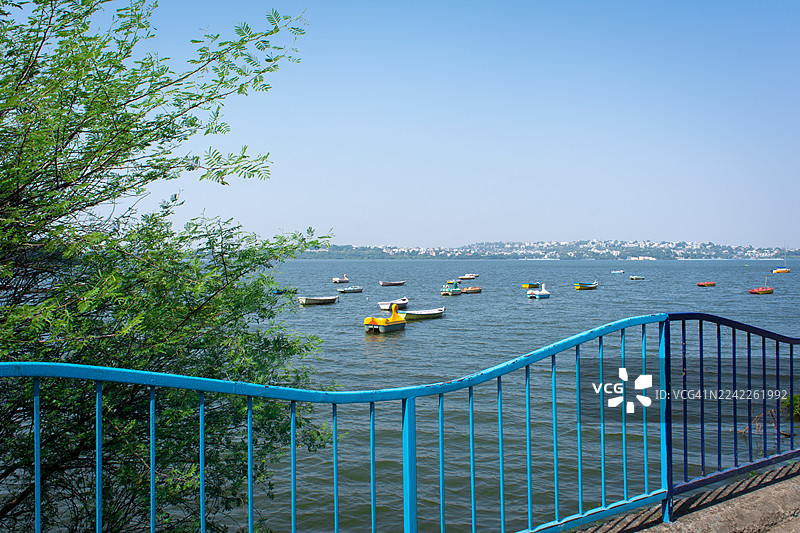 色彩斑斓的脚踏船在湖面上，远处是城市景观，天空湛蓝，被蓝色栏杆和绿色植被环绕，构成一幅风景画。图片素材