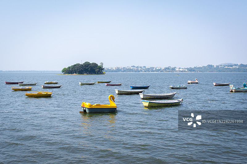 宁静湖泊上停泊着五颜六色脚踏船和划艇，湖中有岛屿，远方是城市天际线，天空晴朗。图片素材