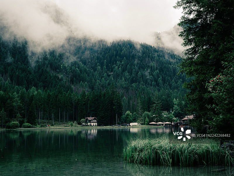 奥地利普斯特里亚山谷，薄雾笼罩的森林湖泊，岸边有一座小屋，周围是茂密的常绿树，天空多云图片素材